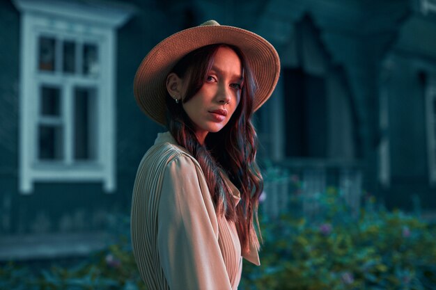 Une fille mystérieuse vêtue d'une robe et d'un chapeau de paille se tient près d'une vieille maison en bois regardant directement dans l'appareil photo une séance photo créative dans des tons rouges photo de haute qualité