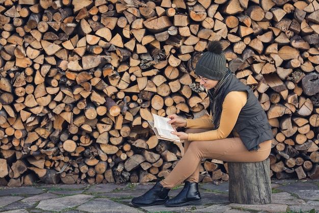 Une fille nerd dans des verres est assise à l'extérieur près d'une pile de bois de chauffage et lit un livre.