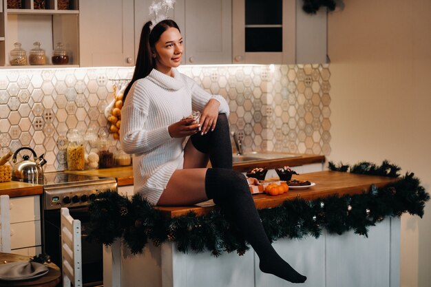 Une fille à Noël est assise sur la table dans la cuisine, tenant un gâteau et rêvant.Une femme le soir du nouvel an dans la cuisine
