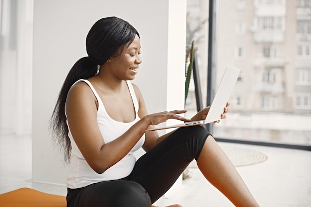 Fille noire assise sur un tapis de yoga orange en studio avec grande fenêtre et utilisant un ordinateur portable