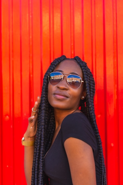 Fille noire de style de vie avec grosse tresse en short en jean tshirt noir et lunettes de soleil sur un mur rouge Portrait d'un jeune danseur de piège