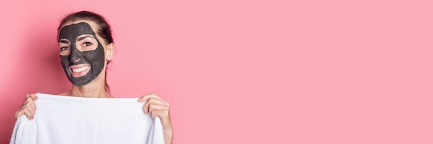Photo fille nue souriante se cachant derrière une serviette avec un masque d'argile sur son visage sur fond rose bannière