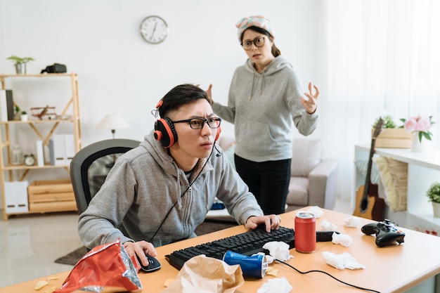 fille offensée debout derrière un petit ami paresseux jouant à un jeu d'ordinateur en ligne. les jeunes couples asiatiques le jour de l'été restent à la maison. petite amie en colère en colère contre un gars concentré en regardant l'écran du pc dans le casque