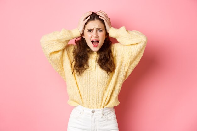 Fille panique et crie à vous jeune femme frustrée se sentir déçu se tenant la main sur la tête et crier en colère debout contre le mur rose