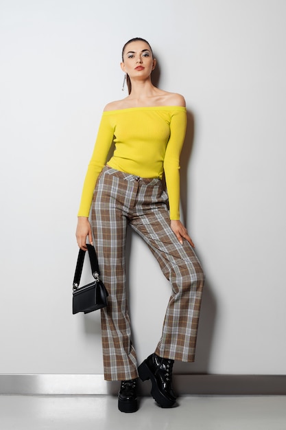 Fille en pantalon à carreaux et pull jaune avec petit sac à main posant près du mur blanc