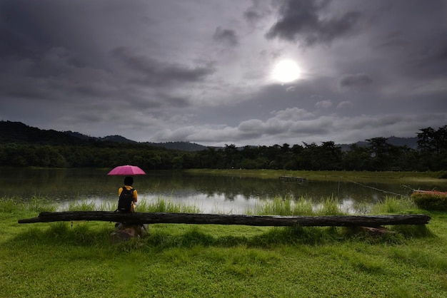 Une fille avec un parapluie rose est assise sur un long siège en bois au bord d'un lac un matin