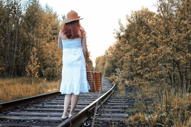 Fille de parc d'automne dans une robe d'été blanche et une valise en osier marchant sur des rails