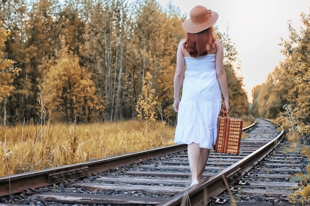 Fille de parc d'automne dans une robe d'été blanche et une valise en osier marchant sur des rails