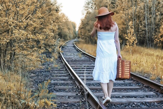 Fille de parc d'automne dans une robe d'été blanche et une valise en osier marchant sur des rails