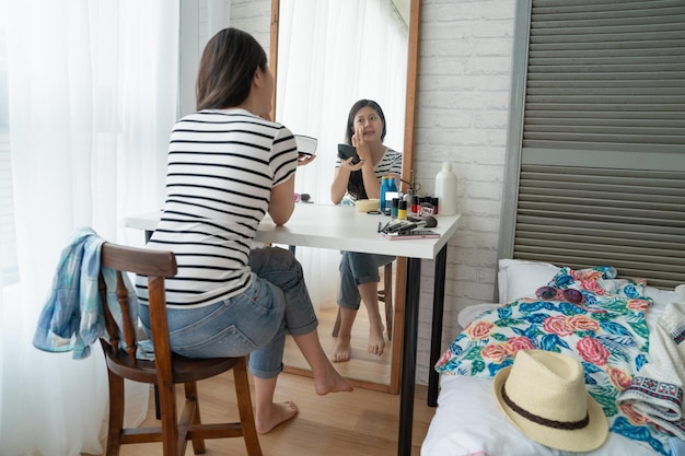 Fille partant en vacances se préparer dans la chambre assis devant le miroir. jeune femme asiatique en tenue décontractée se réveiller tôt en faisant du maquillage dans une coiffeuse se regardant avec des cosmétiques dans un appartement confortable