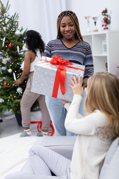 Photo une fille à la peau noire présente un cadeau à son amie pendant les fêtes arbre de noël