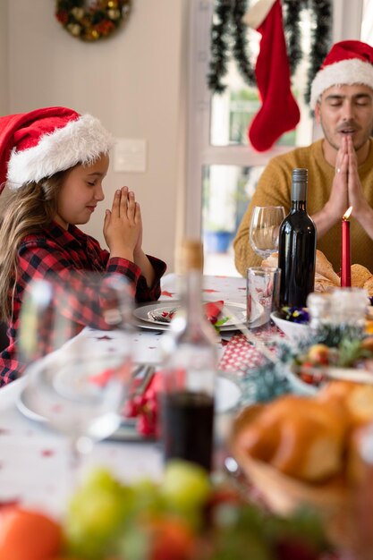 Photo fille et père caucasiens portant des chapeaux de père noël priant à la table de noël. noël en famille et festivités ensemble à la maison.