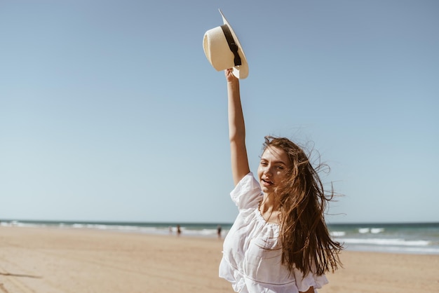 La fille sur la plage a levé son chapeau haut le vent ébouriffe ses cheveux luxueux elle sourit sur le fond du sable de la plage et de l'océan