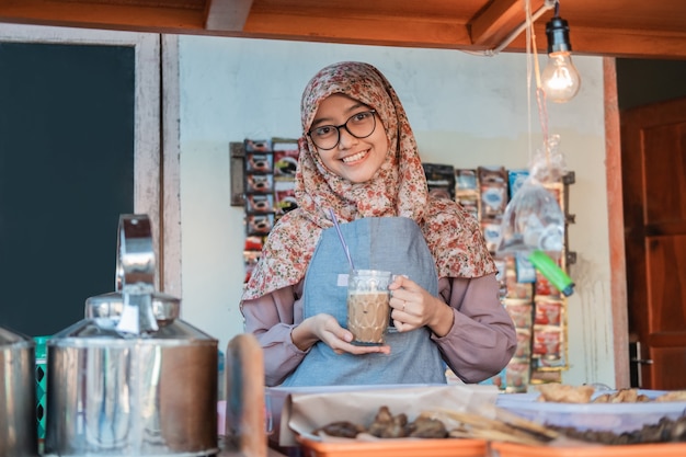 Une fille portant un hijab dans un tablier sourit tout en tenant un verre de café dans un étal de chariot