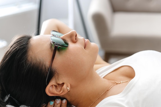 Une fille portant des lunettes de protection pour l'épilation au laser est allongée sur le dos pendant la procédure d'un cosmetol