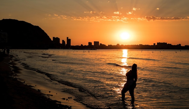 Fille posant contre la lumière au lever du soleil sur la plage de postiguet à Alicante, avec des bâtiments contre