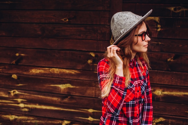 Fille posant dans le contexte de la clôture sur l'ancienne ferme, la vie rurale. fille décontractée, vêtue d'une chemise à carreaux rouge et bleue, d'un jean, de lunettes de soleil et d'un chapeau, avec une clôture en bois marron