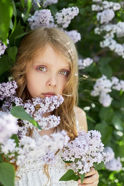 Fille pose dans un buisson de lilas au printemps. Portrait romantique d'un enfant en fleurs à la lumière du soleil