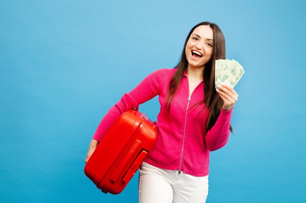 Fille positive avec bagages et argent vont voyager Femme en pull rose et jeans debout sur fond bleu Concept de vacances