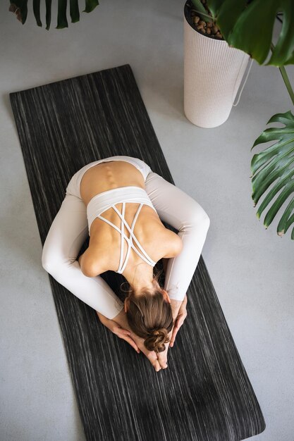 Une fille pratiquant le yoga s'entraîne assise sur un tapis en tenue de sport blanche dans la chambre