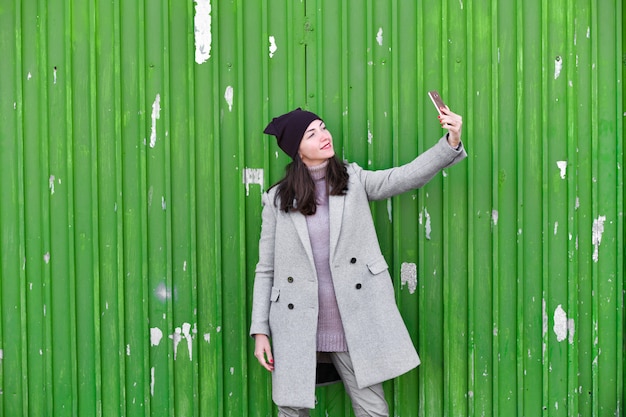 Fille prend un selfie sur une porte industrielle verte. portant un chapeau et un manteau. lieu d'écriture. vêtu d'une pagode froide. vêtements et style. émotions naturelles