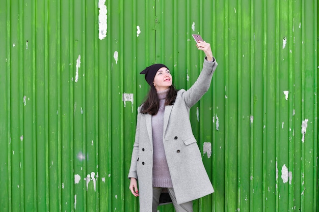 Fille prend un selfie sur une porte industrielle verte. portant un chapeau et un manteau. lieu d'écriture. vêtu d'une pagode froide. vêtements et style. émotions naturelles