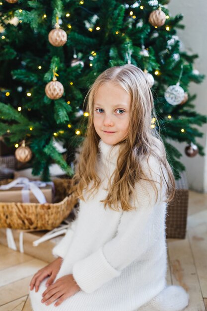 Fille près de l'arbre de Noël décoré de boules et de guirlandes, fille aux cheveux longs dans un pull blanc, en attendant le nouvel an et Noël, cadeaux