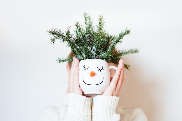 Une fille en pull blanc cache sa tête derrière une tasse avec un visage de bonhomme de neige et des branches de sapin.