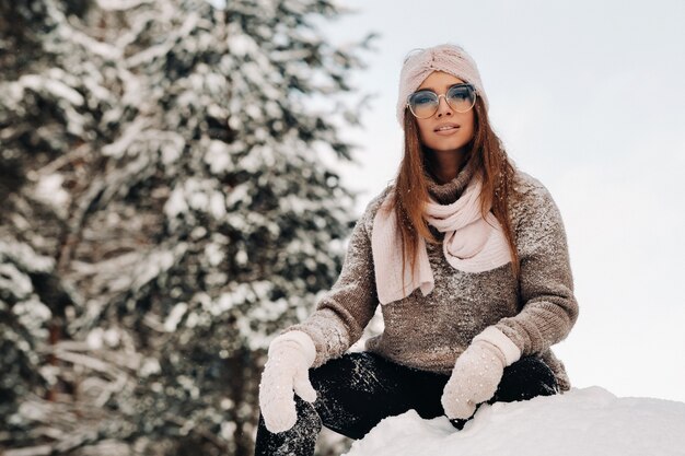 Une fille en pull et lunettes en hiver est assise sur un fond enneigé dans la forêt.