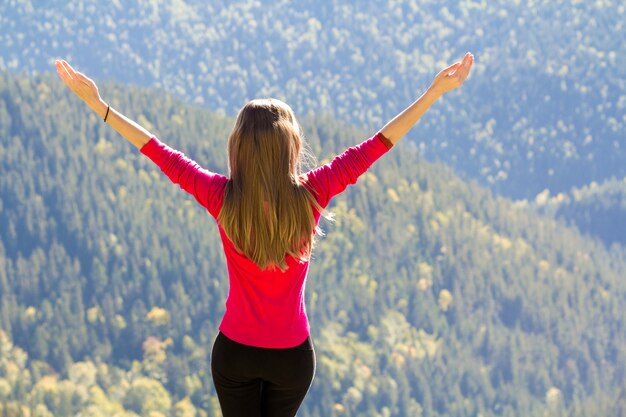 Fille en pull rouge debout sur de gros rochers dans les montagnes en levant les mains.