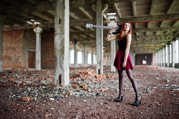 Fille punk aux cheveux rouges portant une jupe noire et rouge avec une guitare basse à un endroit abandonné Portrait d'une musicienne gothique