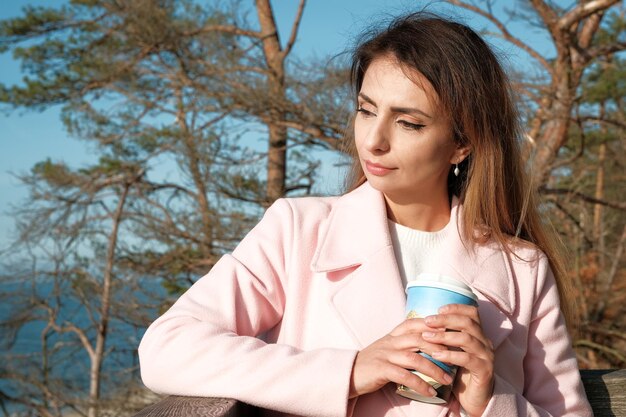 Fille qui marche le long du bord de la mer brune boit du café fille dans un manteau rose