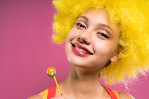 Photo fille qui rit dans une perruque orange vif sur fond rose tient dans les mains des chupa chups