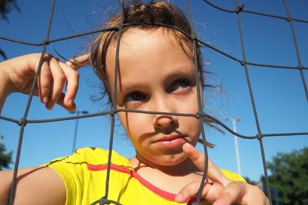 Une fille de race blanche de 8 ans se tient derrière un filet de sport et lève les yeux pensivement Grimace sur le visage Sports de volley-ball divisant le filet contre le ciel bleu gros plan Sports de plein air