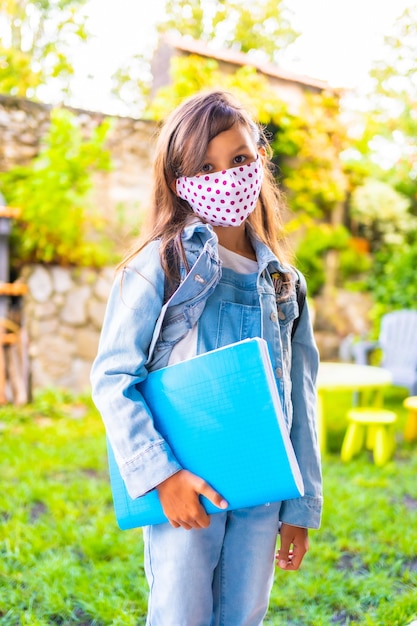 Fille de race blanche avec un masque facial prêt à retourner à l'école. Nouvelle normalité, distance sociale, pandémie de coronavirus, covid-19. Veste, sac à dos, masque à pois roses et bloc bleu à la main