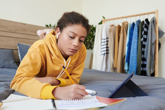 Fille de race mixte ciblée dans les écouteurs allongée sur le lit et utilisant une tablette numérique tout en faisant hometask