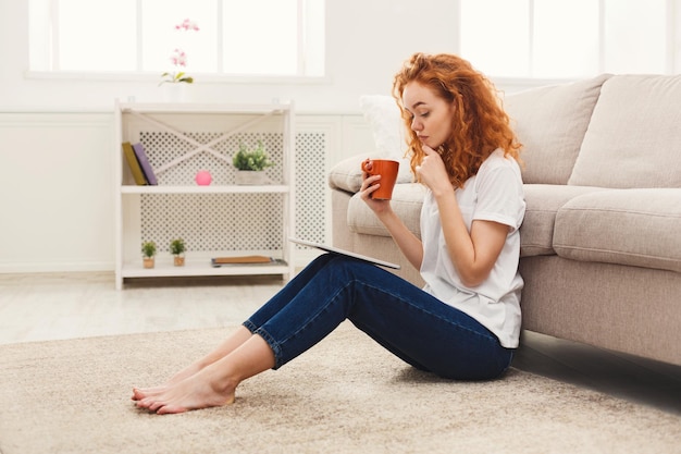 Fille réfléchie lisant sur tablette. Jeune femme rousse décontractée utilisant un appareil numérique assis sur le sol à la maison, espace pour copie