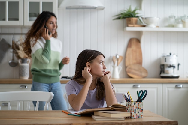 Une fille réfléchie et non concentrée regarde de côté pour faire ses devoirs est assise à table avec des manuels à la cuisine