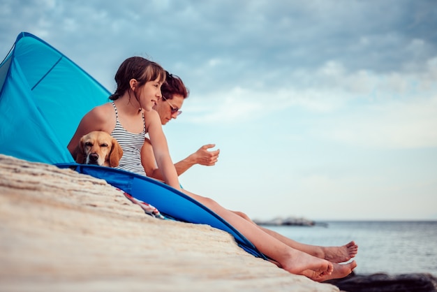 Photo fille regardant au coucher du soleil assis dans une tente de plage avec sa mère et son chien