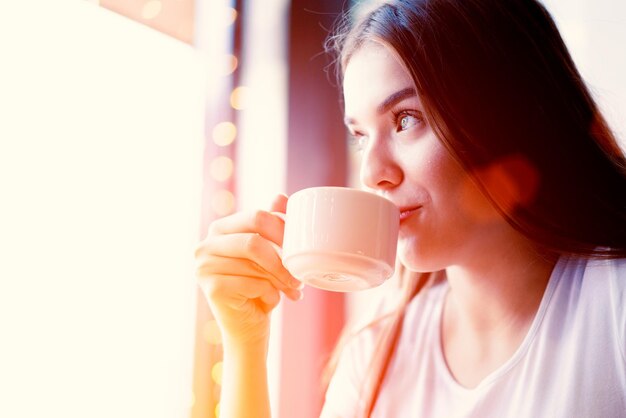 Fille regardant par la fenêtre en buvant du café