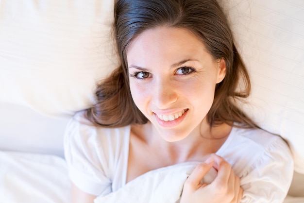 Fille rêveuse allongée sur un lit blanc avec de grands oreillers étreignant une couverture avec elle