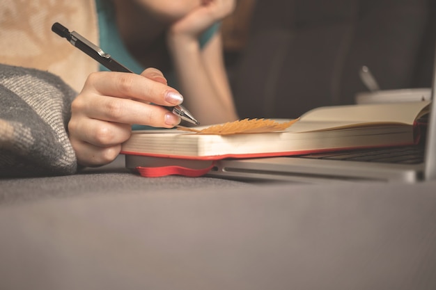 Une fille rêveuse écrit un journal à la maison pendant les vacances d'automne, assise sur un canapé avec un plaid chaud. main avec un crayon et des feuilles d'automne sur le livre