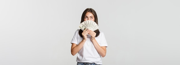 Fille rêveuse excitée tenant de l'argent et regardant le coin supérieur gauche pensant sur fond blanc