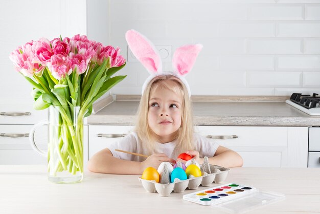 Une fille rêveuse avec des oreilles de lapin peint des oeufs de pâques à une table en bois