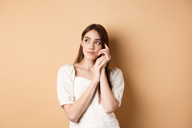 Fille rêveuse et romantique à la recherche de côté avec un regard pensif, touchant la peau du visage douce et propre, debout sur un fond beige.