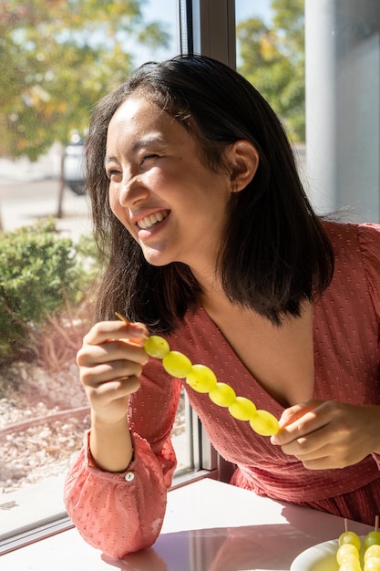 Fille riant devant la fenêtre recevant la lumière du soleil tenant une brochette de raisins