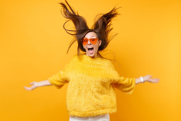 Fille de rire en pull de fourrure et lunettes orange coeur s'amuser en studio avec des cheveux volants isolés sur fond jaune vif. Les gens émotions sincères, concept de style de vie. Espace publicitaire.