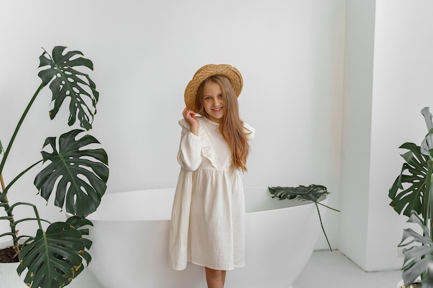 une fille en robe blanche et un chapeau de paille dans une chambre avec des plantes et une salle de bain