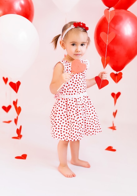 Fille En Robe Blanche Avec Des Coeurs Mangeant Une Sucette En Forme De Coeur. Bébé Tient Des Boules Rouges Et Blanches Pour La Saint-valentin