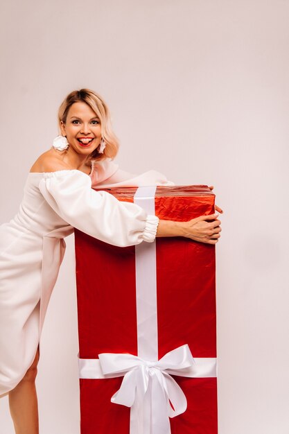 Une fille en robe blanche avec un énorme cadeau rouge sur fond blanc sourit.